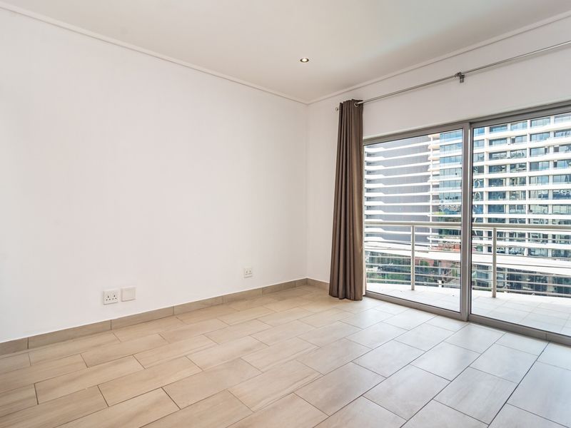 A bright, empty bedroom featuring modern tiled flooring, recessed lighting, and a large sliding glass door providing access to a private balcony.
