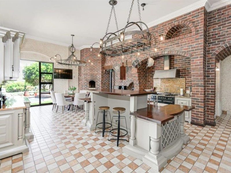 Spacious open-plan kitchen featuring a large island, brick feature wall with built-in oven and pizza oven, and elegant chandeliers.