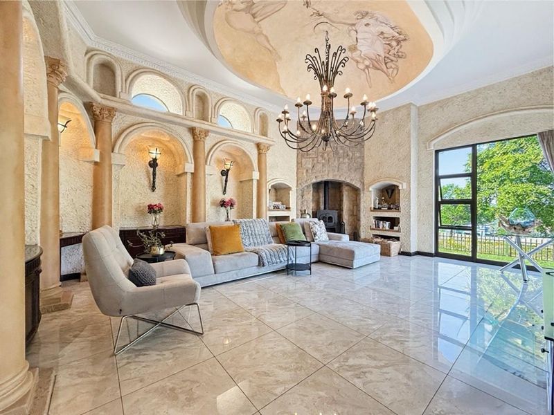 Spacious living room featuring a decorative ceiling mural, stone fireplace, classical architectural columns, built-in wall niches, and polished marble-look floor tiles.