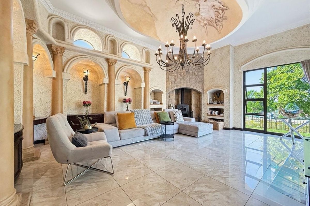 Spacious living room featuring a decorative ceiling mural, stone fireplace, classical architectural columns, built-in wall niches, and polished marble-look floor tiles.
