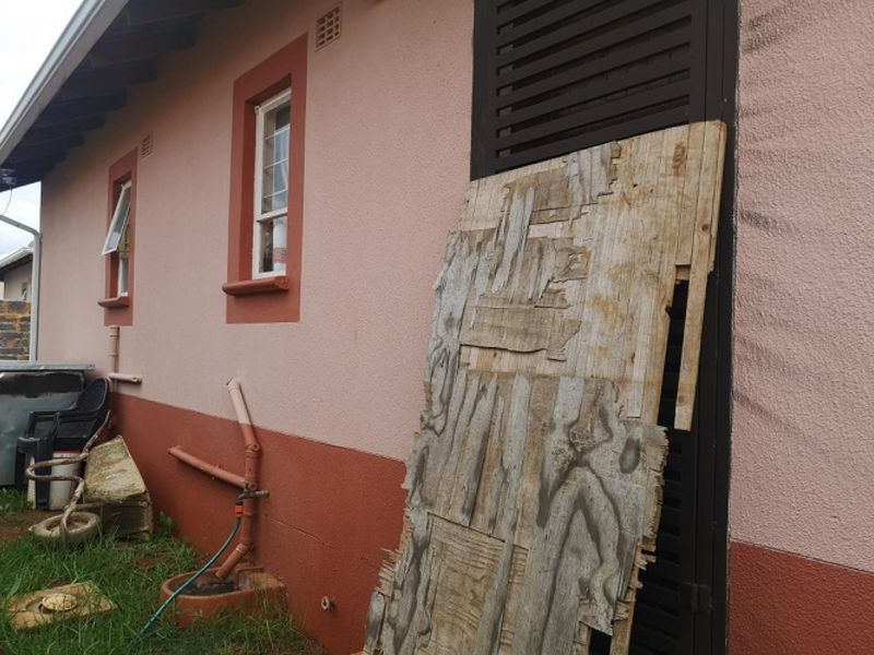 Rear exterior view of the house showing a damaged door covering, exposed plumbing, and painted masonry walls.