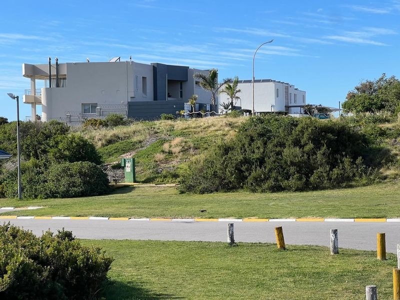 A view of the residential street frontage showing the vacant land plot with established grass, utility infrastructure, and neighbouring properties in the background.