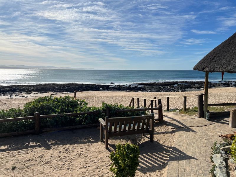 Scenic coastal view overlooking the beach and ocean, featuring a thatched roof structure and paved pathway access.