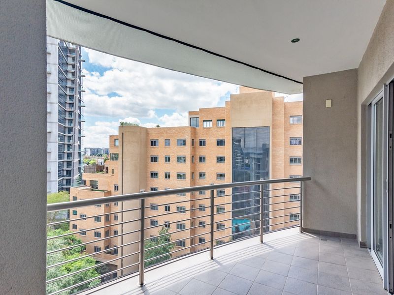 A clean, covered balcony featuring tiled flooring, a modern stainless steel balustrade, and recessed lighting, offering an urban view of surrounding apartment buildings.