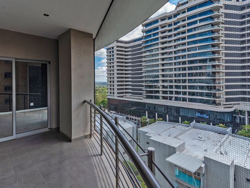 A spacious covered balcony featuring tiled flooring and a modern stainless steel balustrade, offering an elevated urban view of the surrounding apartment complex.