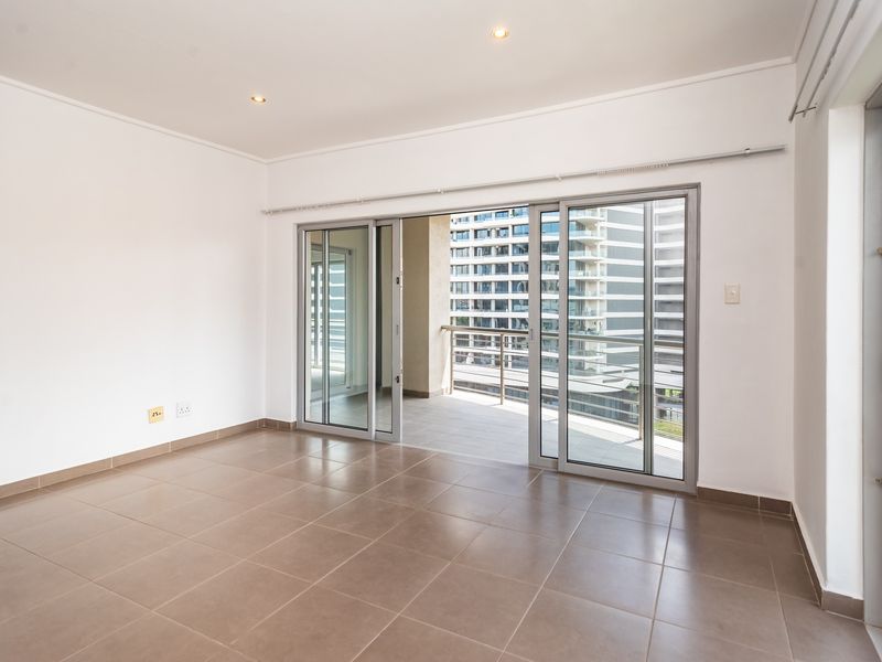 An empty, well-lit living area featuring neutral tiled flooring and large sliding glass doors that open onto a private balcony.
