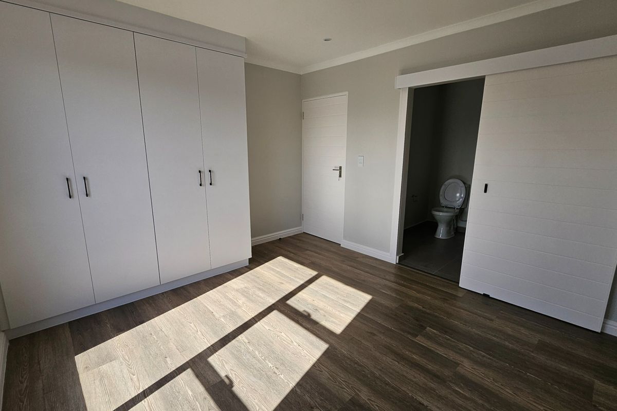 A bright, modern bedroom featuring built-in cupboards, durable laminate flooring, and a convenient en-suite toilet accessed via a sliding door.