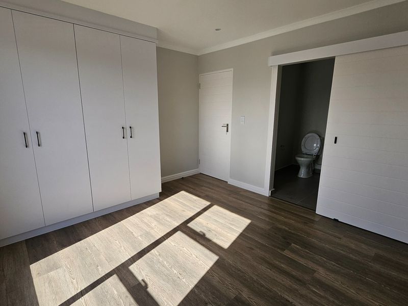 A bright, modern bedroom featuring built-in cupboards, durable laminate flooring, and a convenient en-suite toilet accessed via a sliding door.