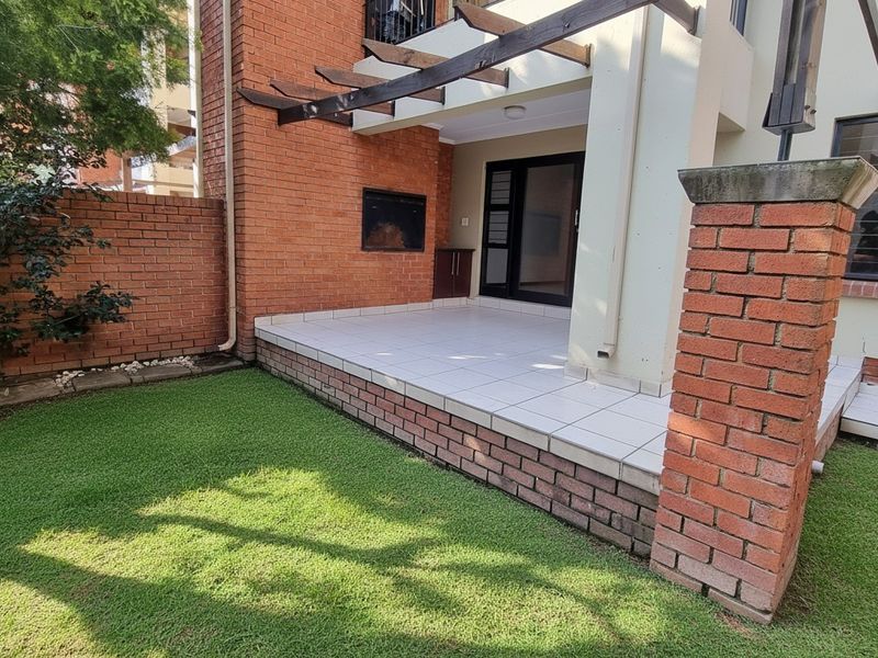 A private patio area featuring a built-in braai, tiled flooring, and a timber pergola, overlooking a small garden space.