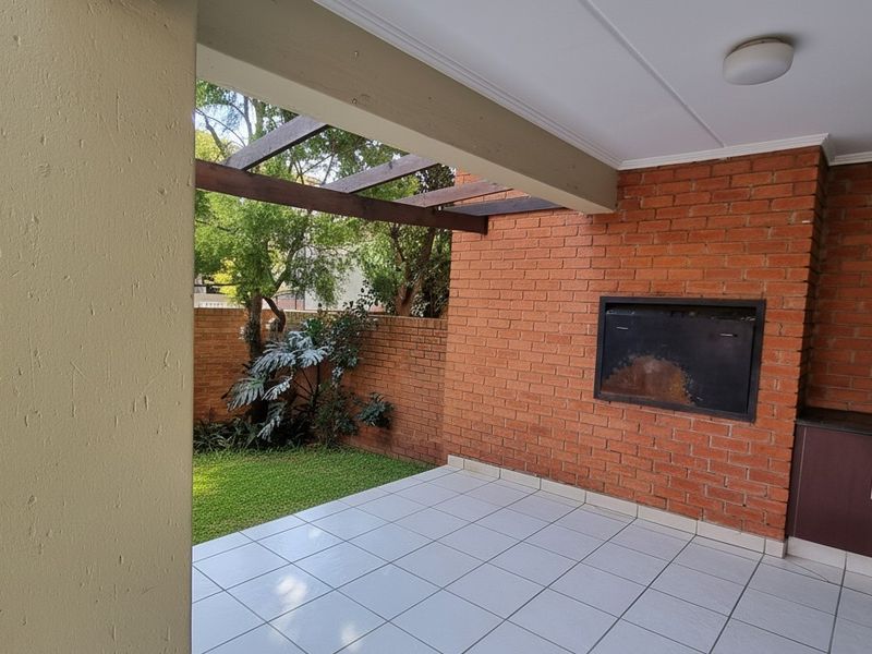 Covered patio area featuring a built-in braai, tiled flooring, and an exposed timber pergola, overlooking a small private garden space.