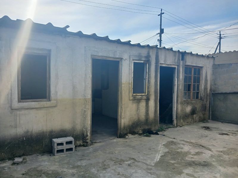 Rear exterior view of a dwelling showing significant wear to the plaster and paint, with multiple window frames and concrete patio area requiring maintenance.