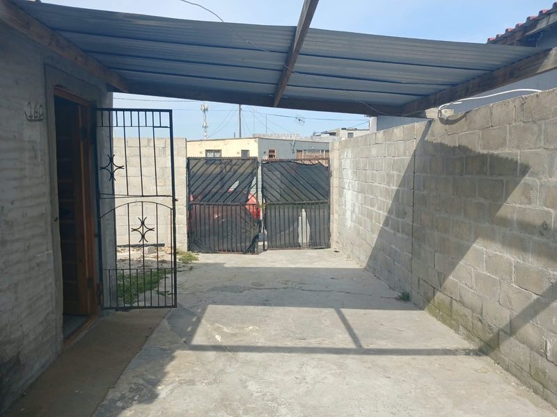 A functional front entrance area featuring a concrete driveway, secure metal gate, and a covered parking space with a corrugated metal roof.