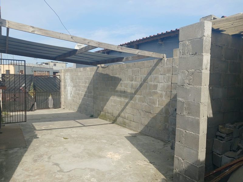 An outdoor area featuring a concrete block boundary wall, metal gate, and a timber pergola frame over a concrete slab.