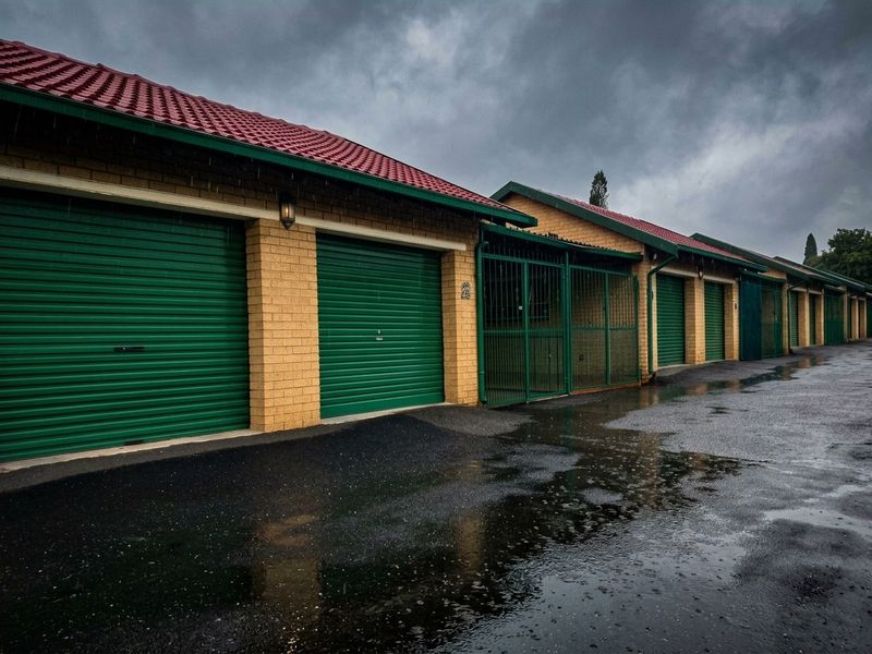 A row of secure garage units featuring roller shutter doors, face brick construction, and a paved driveway under a cloudy sky.