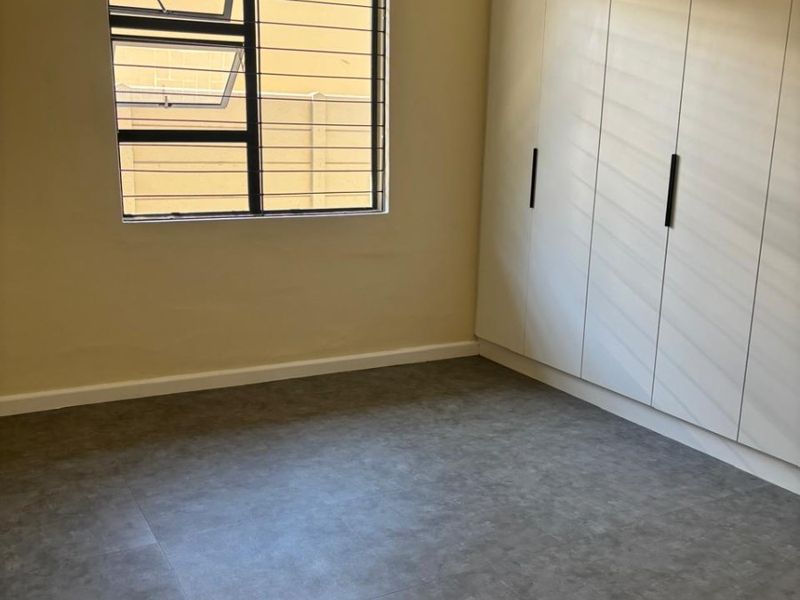 A clean, empty bedroom featuring modern grey vinyl flooring, built-in white cupboards, and a window fitted with security bars.