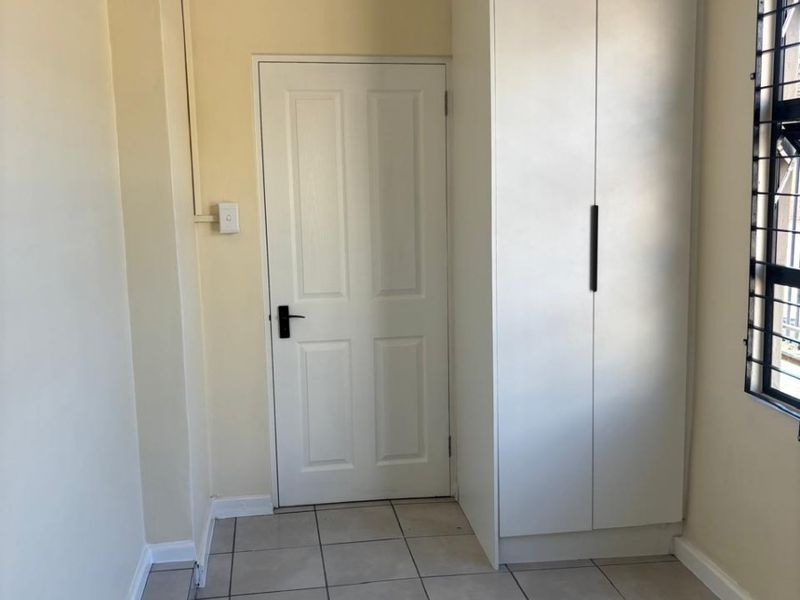 A clean, empty bedroom featuring neutral tiled flooring, a built-in wardrobe, and a window fitted with security bars.