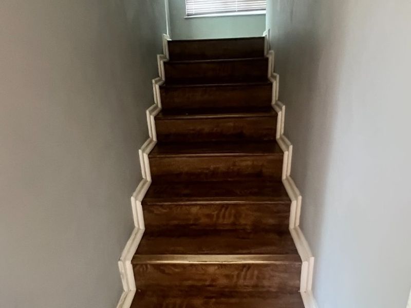 A narrow interior staircase with dark wood-look treads and light-colored walls, leading to an upper landing with a window.