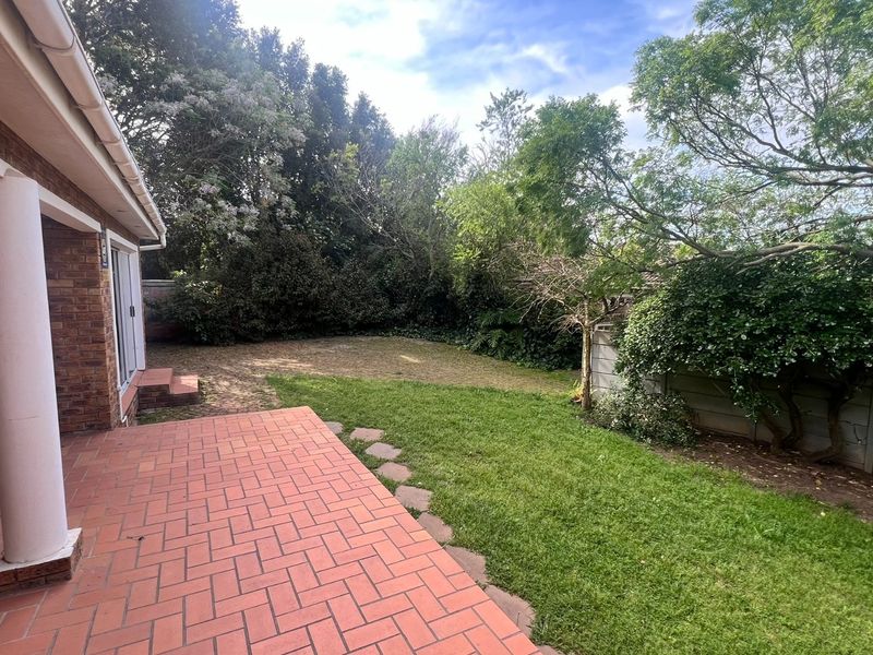 A private rear garden featuring a paved brick patio, covered veranda area, and established lawn with mature trees and boundary walling.