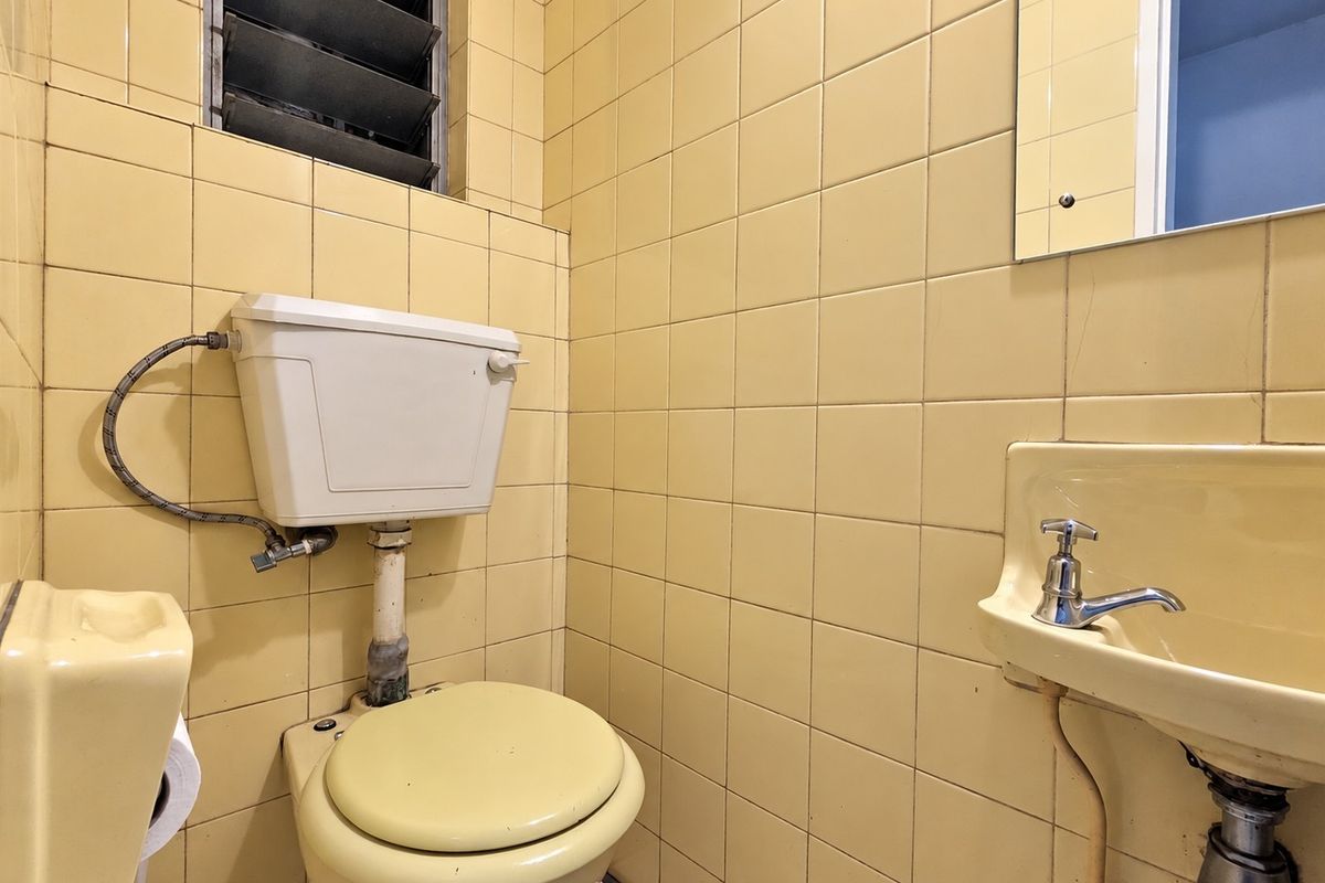 A compact bathroom featuring original yellow wall tiles, a wall-mounted basin, and a louvered window, offering potential for a modern renovation.