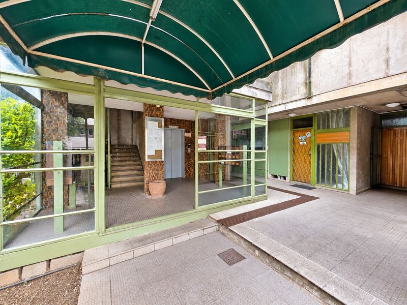 Entrance area of an apartment building featuring a green metal awning, glass-enclosed lobby, elevator access, and a staircase.