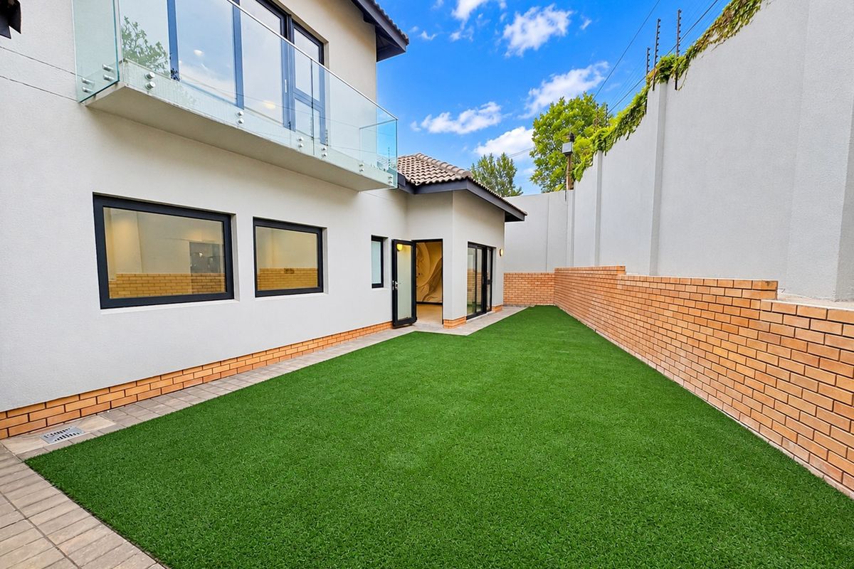 A well-maintained rear garden featuring artificial turf, a brick boundary wall with electric fencing, and a modern upper-level balcony with a glass balustrade.
