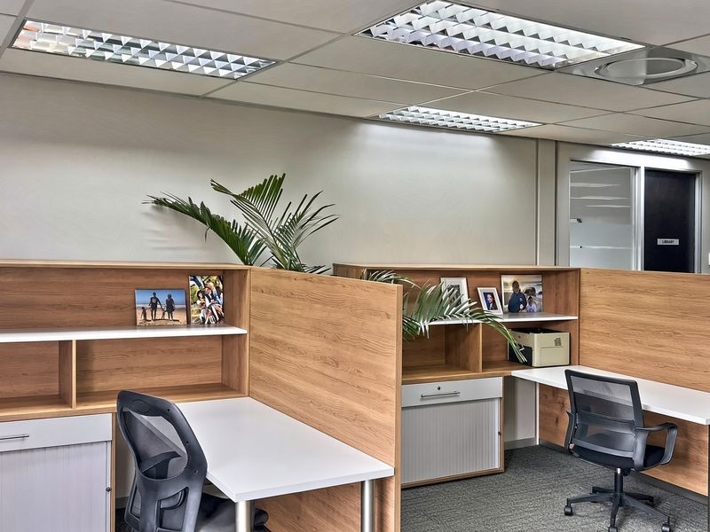 Professional commercial office space featuring built-in wooden cubicle workstations, suspended ceiling grid with recessed lighting, and durable carpet flooring.