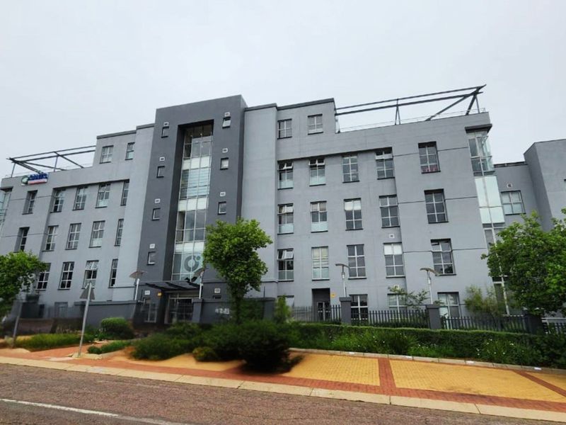 A modern multi-story commercial office building featuring a grey facade, glass architectural elements, and a paved pedestrian entrance area.