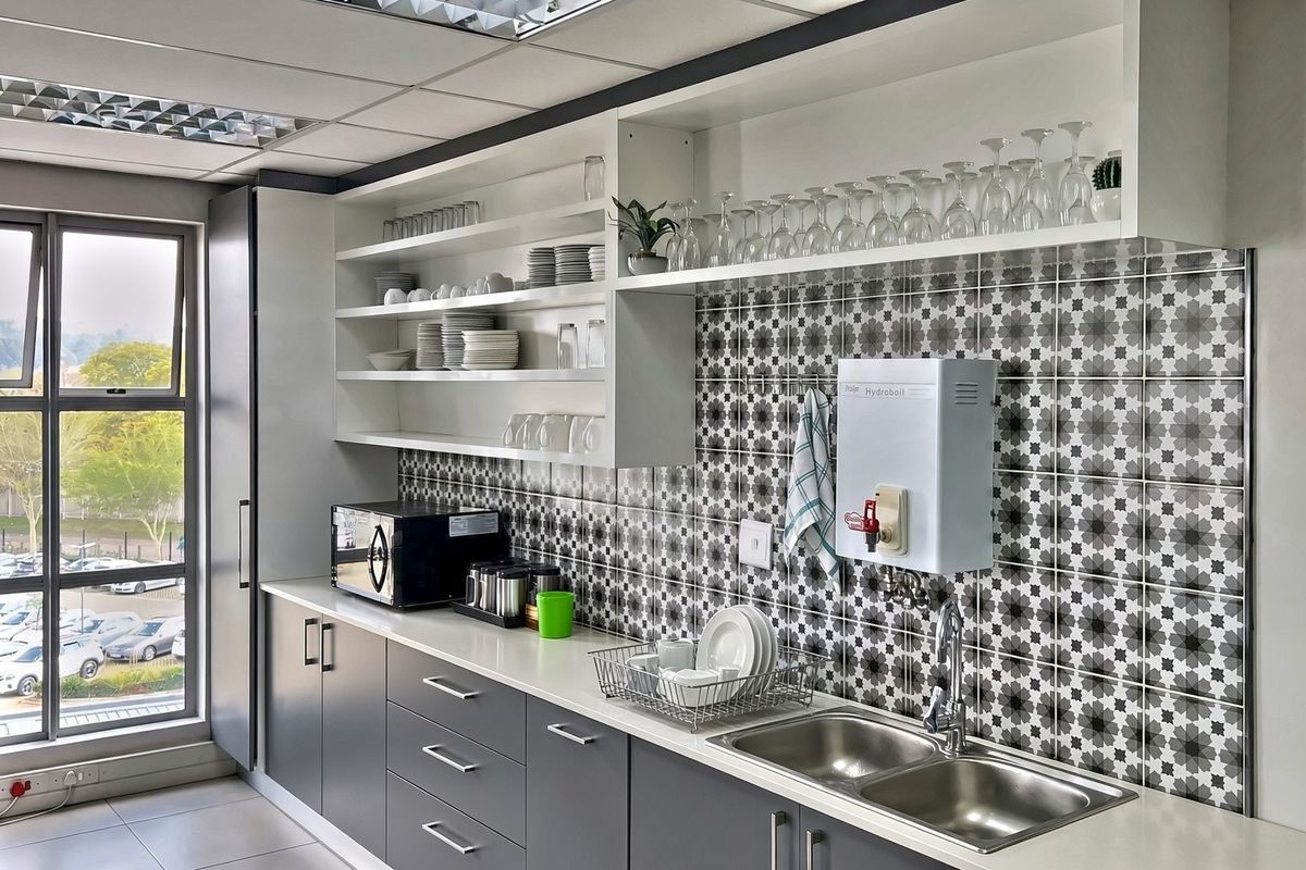 Modern commercial kitchenette featuring grey cabinetry, stainless steel double sink, patterned tiled backsplash, and a wall-mounted hydroboil unit with ample open shelving.