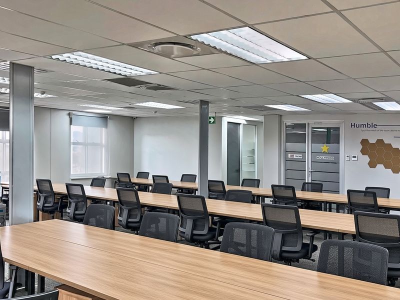 A spacious commercial office area featuring a suspended ceiling, recessed lighting, and carpeted flooring, configured with multiple workstations in an open-plan layout.