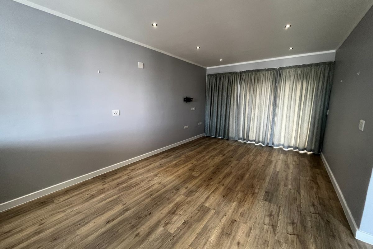 A spacious, empty living room featuring modern laminate wood flooring, recessed downlighting, and large windows covered by curtains.