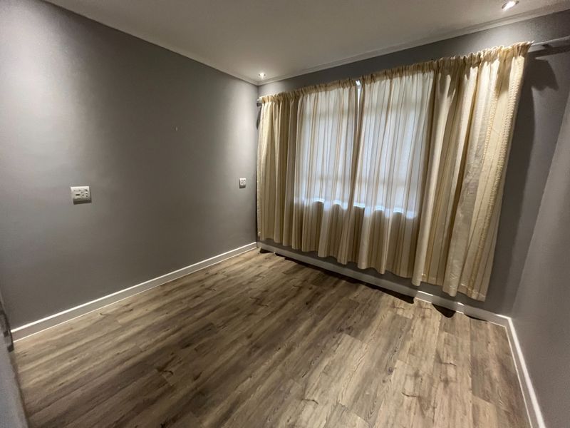 A bright, empty bedroom featuring modern laminate wood-look flooring, neutral grey walls, and recessed ceiling lighting.