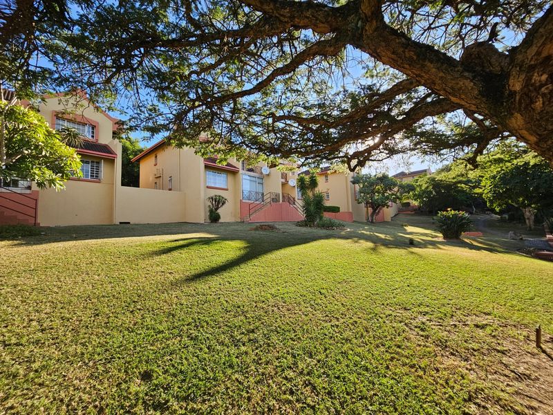 A view of the apartment complex rear exterior featuring a well-maintained lawn, mature trees, and building facades with balconies.