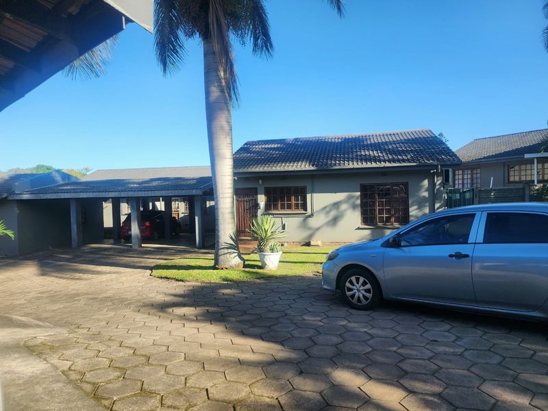 Front exterior view of the townhouse featuring a paved driveway, covered carport, and established garden with a large palm tree.