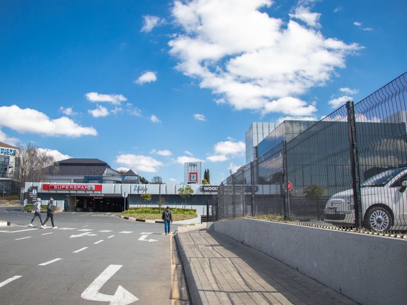 Exterior view of a commercial shopping center entrance featuring paved walkways, security fencing, and access to underground parking facilities.