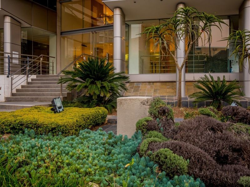 Modern commercial building entrance featuring a glass facade, metal structural columns, and a well-maintained landscaped garden area with concrete stairs.