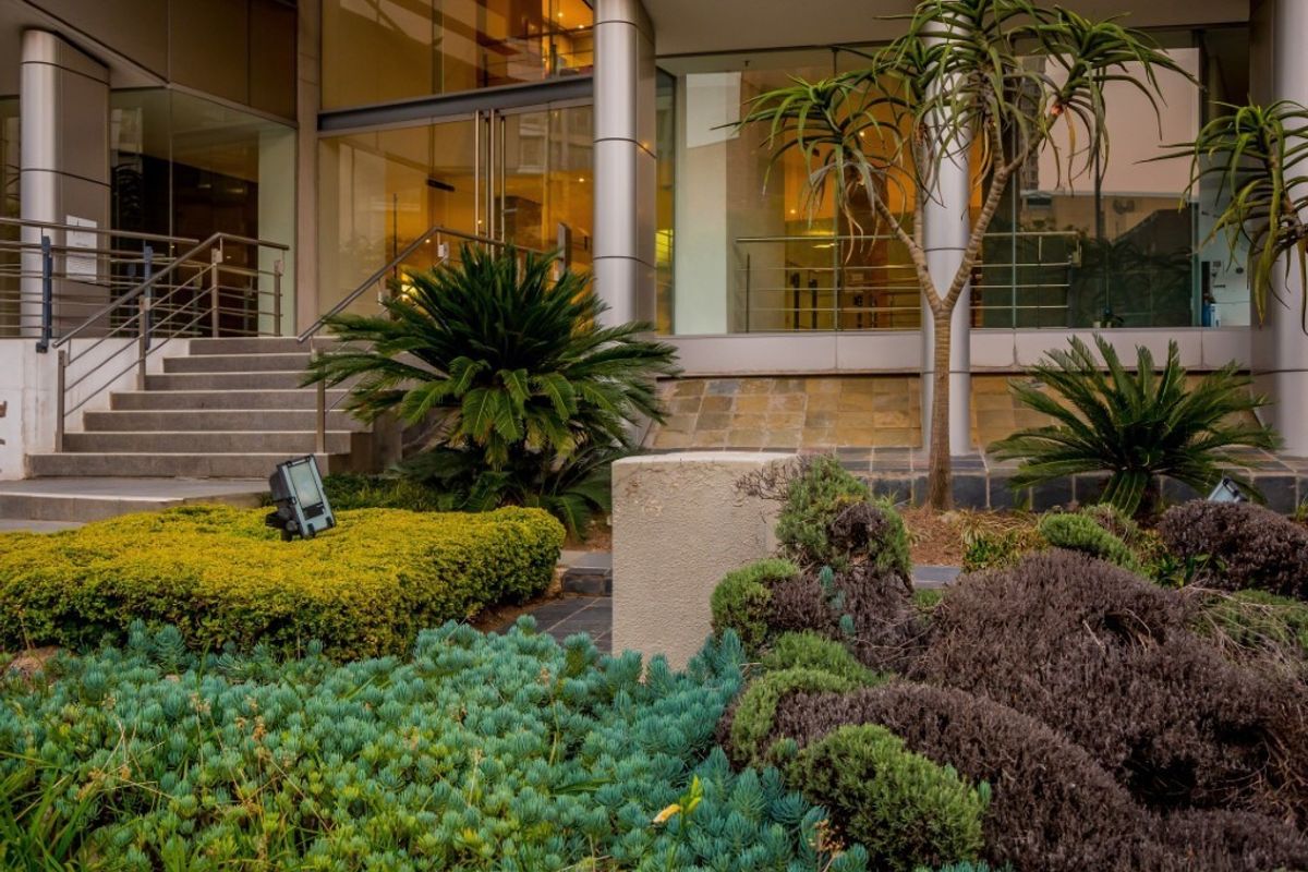 Modern commercial building entrance featuring a glass facade, metal structural columns, and a well-maintained landscaped garden area with concrete stairs.
