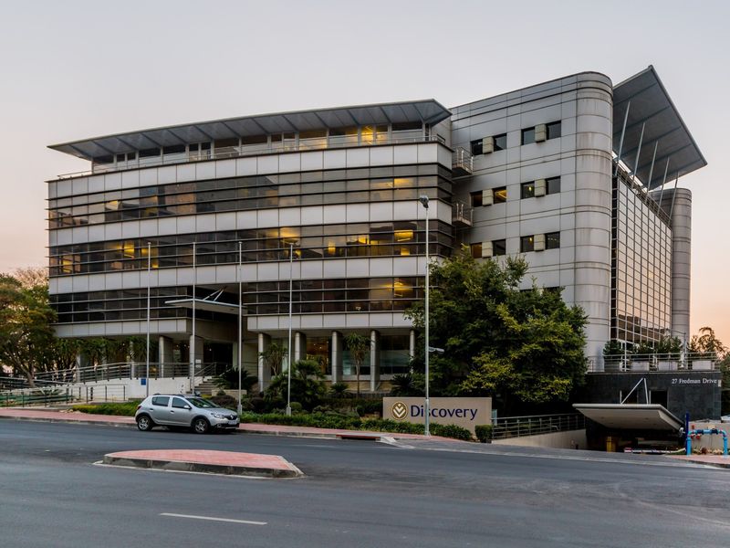 A professional exterior view of a modern multi-story commercial office building featuring glass curtain walls, metal cladding, and a dedicated underground parking entrance.