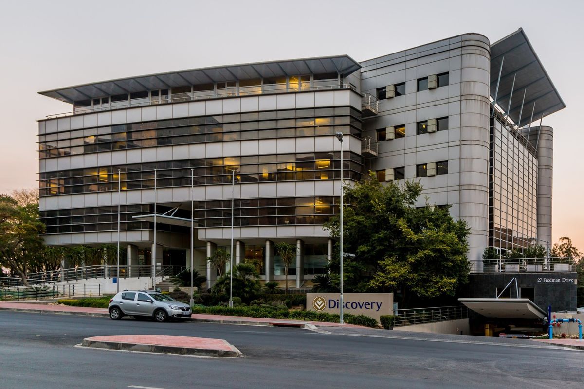 A professional exterior view of a modern multi-story commercial office building featuring glass curtain walls, metal cladding, and a dedicated underground parking entrance.