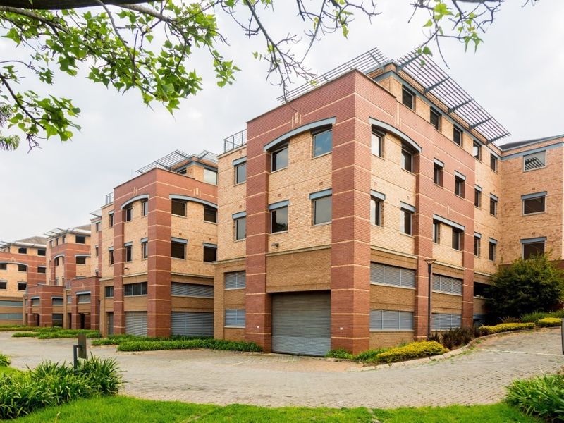 A multi-story commercial office building featuring a face brick facade, paved driveway access, and secure roller shutter doors at the ground level.