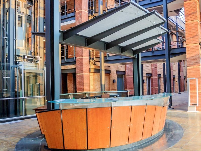 Modern commercial reception area featuring a curved desk, glass elevator, and industrial-style brick and steel architecture within an open-air atrium.