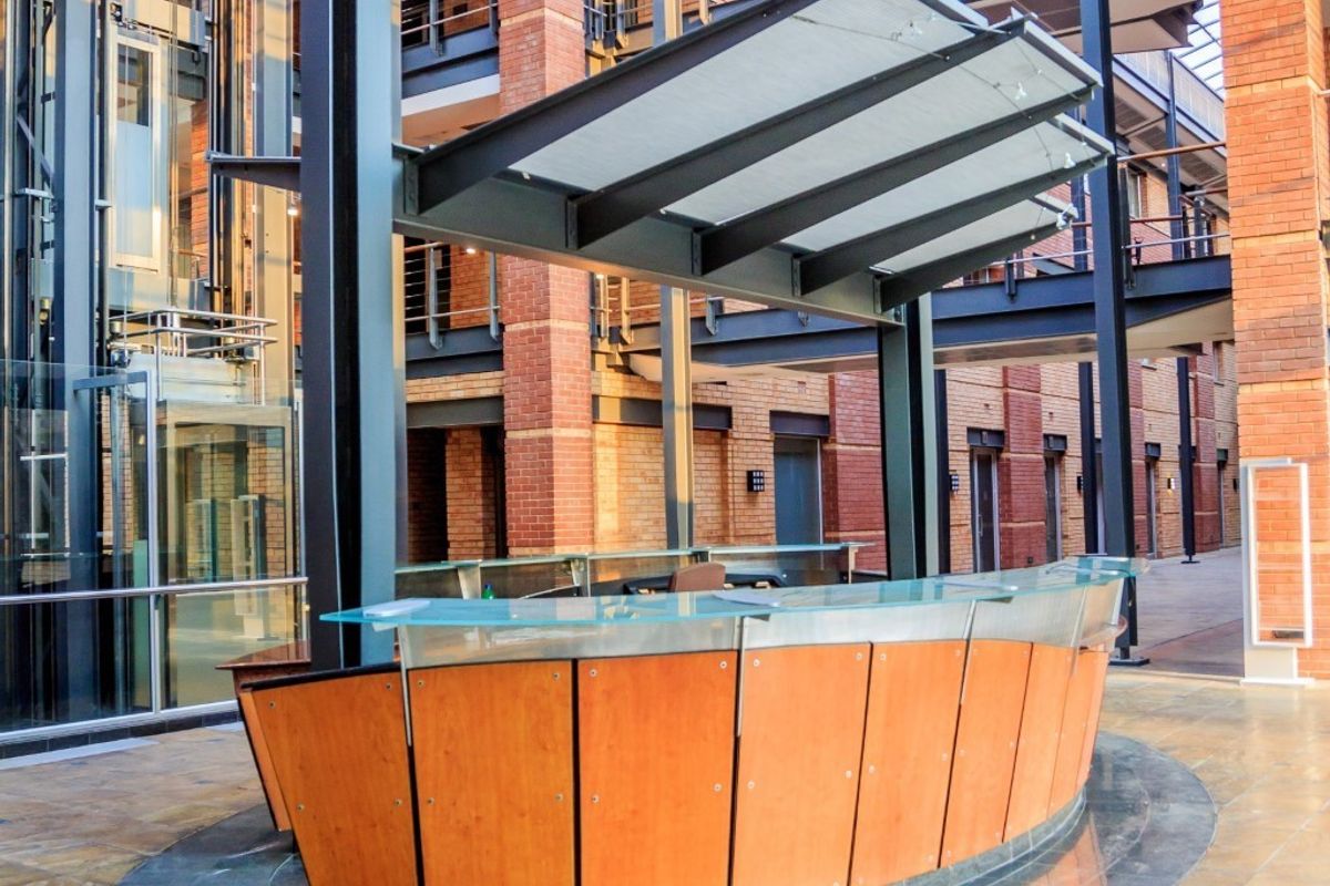Modern commercial reception area featuring a curved desk, glass elevator, and industrial-style brick and steel architecture within an open-air atrium.