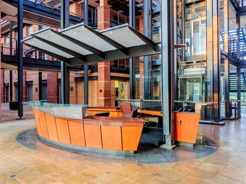 Modern commercial lobby featuring a central reception desk, glass elevator, exposed brick architecture, and industrial steel structural elements.