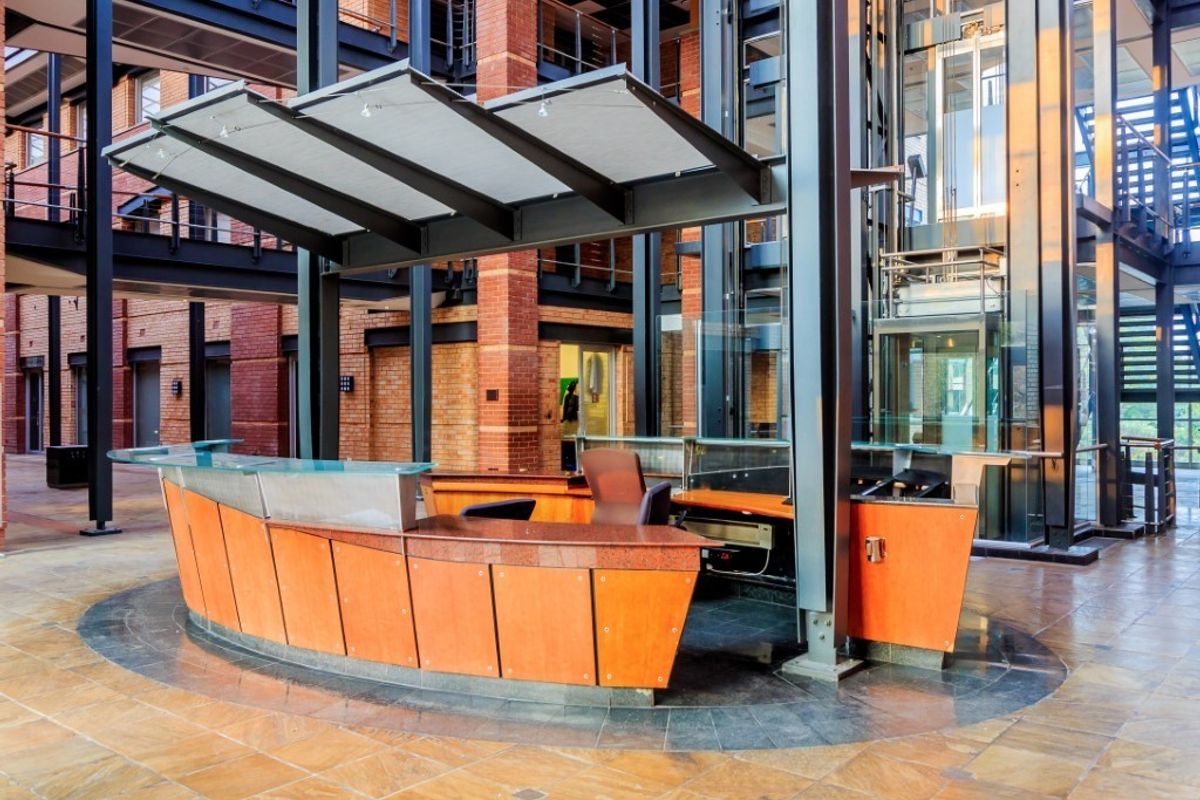 Modern commercial lobby featuring a central reception desk, glass elevator, exposed brick architecture, and industrial steel structural elements.