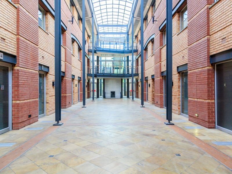 A spacious, modern commercial atrium featuring a high glass roof, exposed brick walls, and stone flooring, providing a bright and professional entrance area.