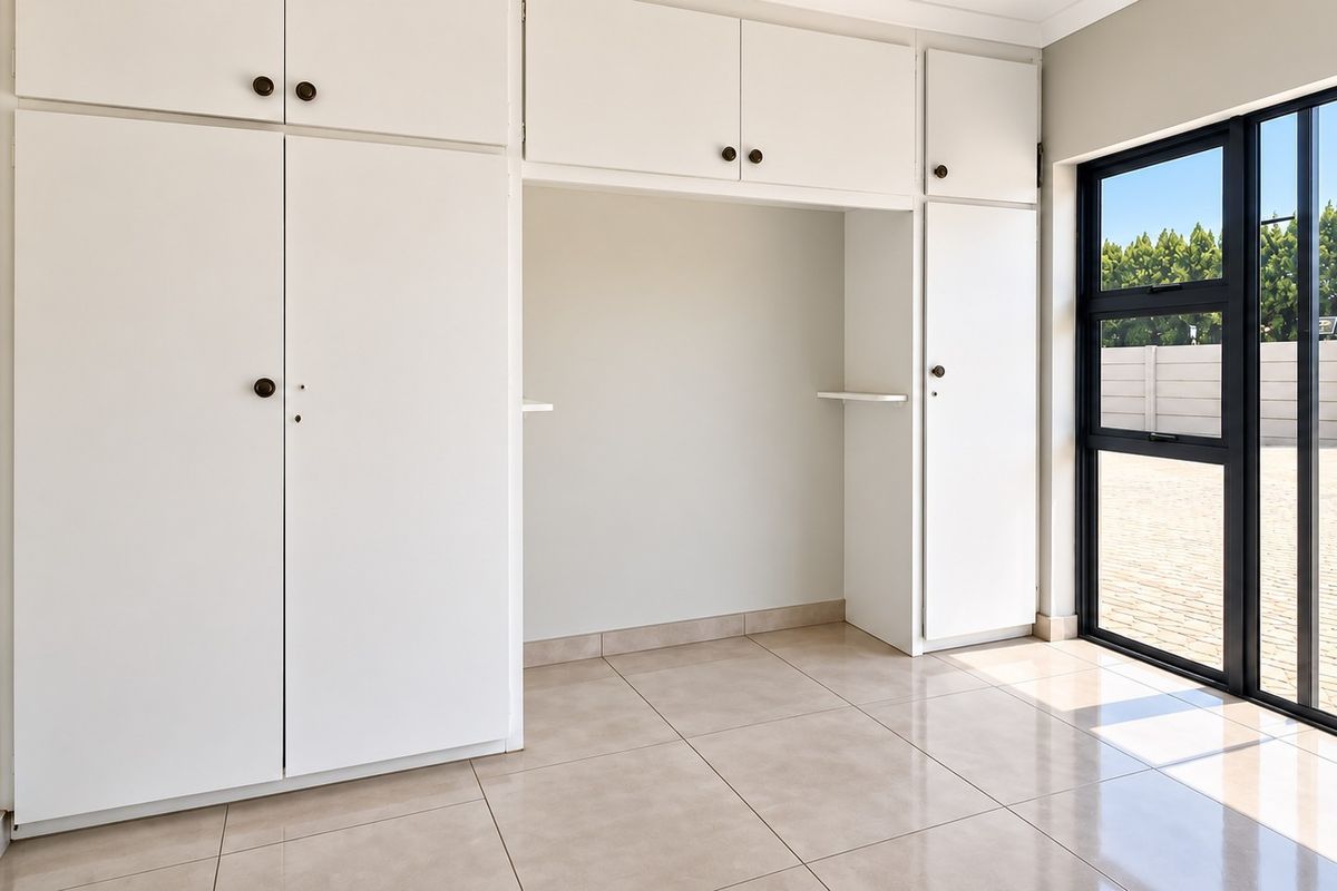 A bright, empty bedroom featuring extensive built-in white cabinetry, neutral tiled flooring, and large aluminium-framed windows providing natural light.