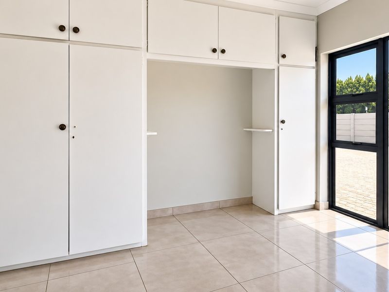 A bright, empty bedroom featuring extensive built-in white cabinetry, neutral tiled flooring, and large aluminium-framed windows providing natural light.
