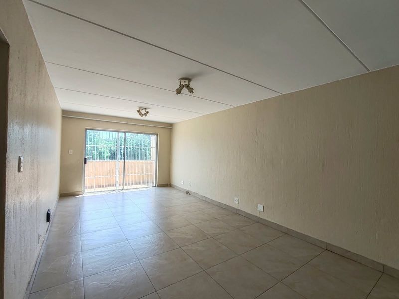 A spacious, empty living area featuring neutral tiled flooring, ceiling-mounted lighting, and a sliding glass door leading to an outdoor balcony with security gate.