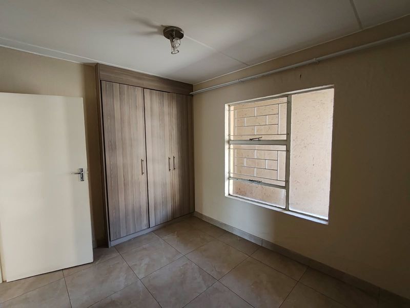 A clean, empty bedroom featuring tiled flooring, a built-in wooden wardrobe, and a window fitted with security bars.
