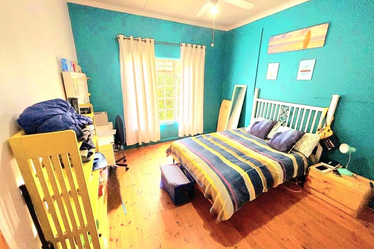 A bedroom featuring timber flooring, a ceiling fan, and a window with curtains.