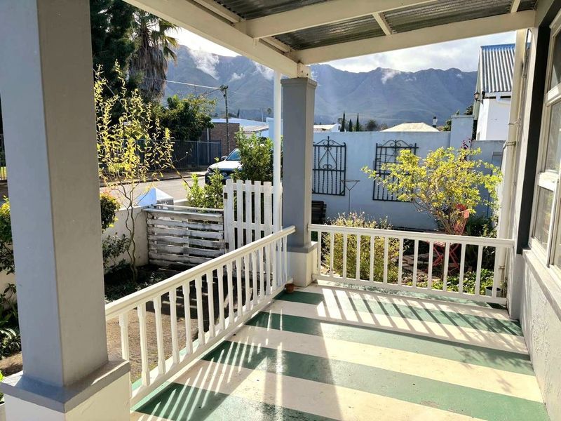 A covered front patio featuring a painted striped floor, support columns, and a white railing, offering a view of the surrounding garden and mountains.